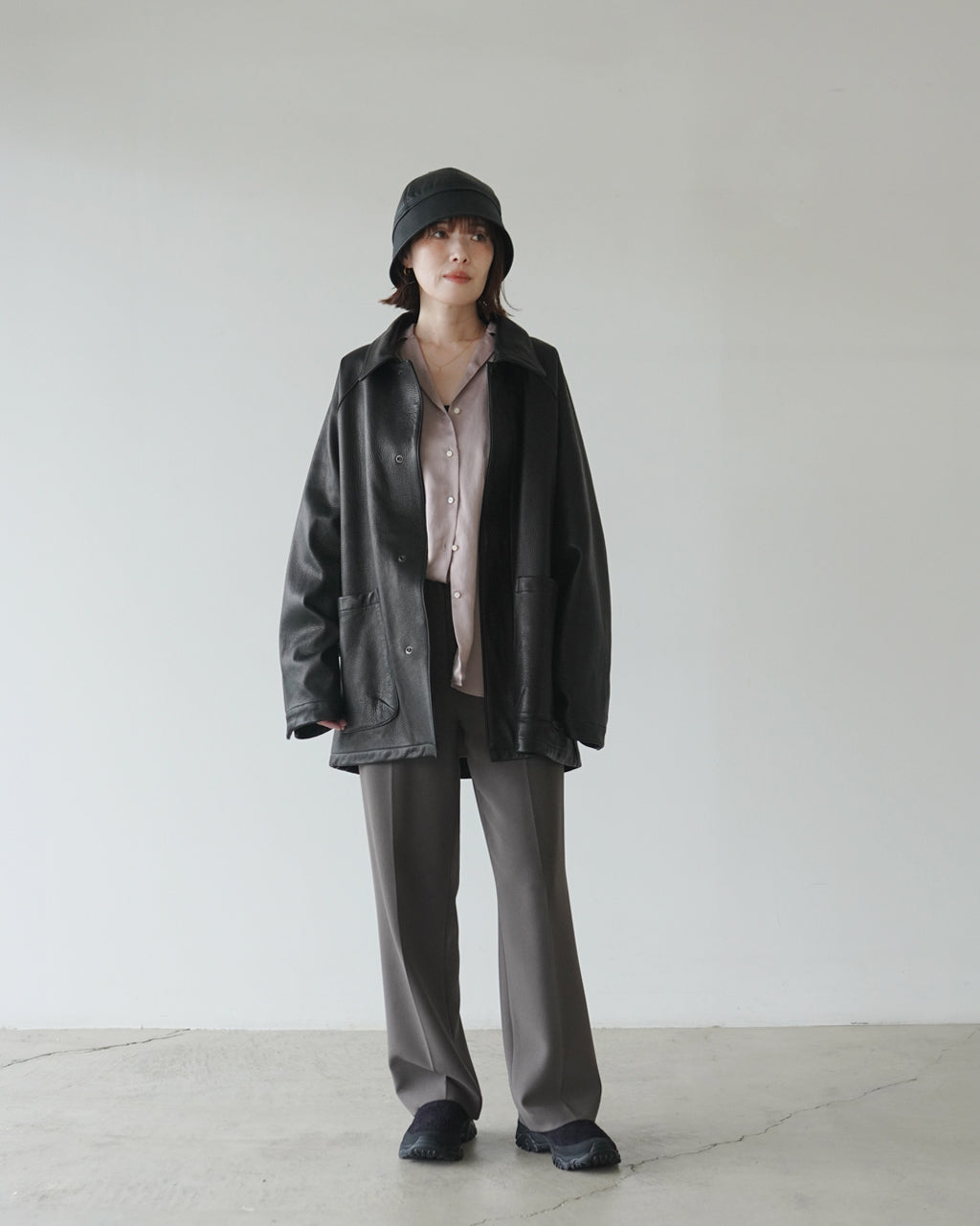 黒のレザージャケットとベージュのシャツ、くすんだグレーパンツを着用した女性が帽子をかぶり正面へ立つ全身写真