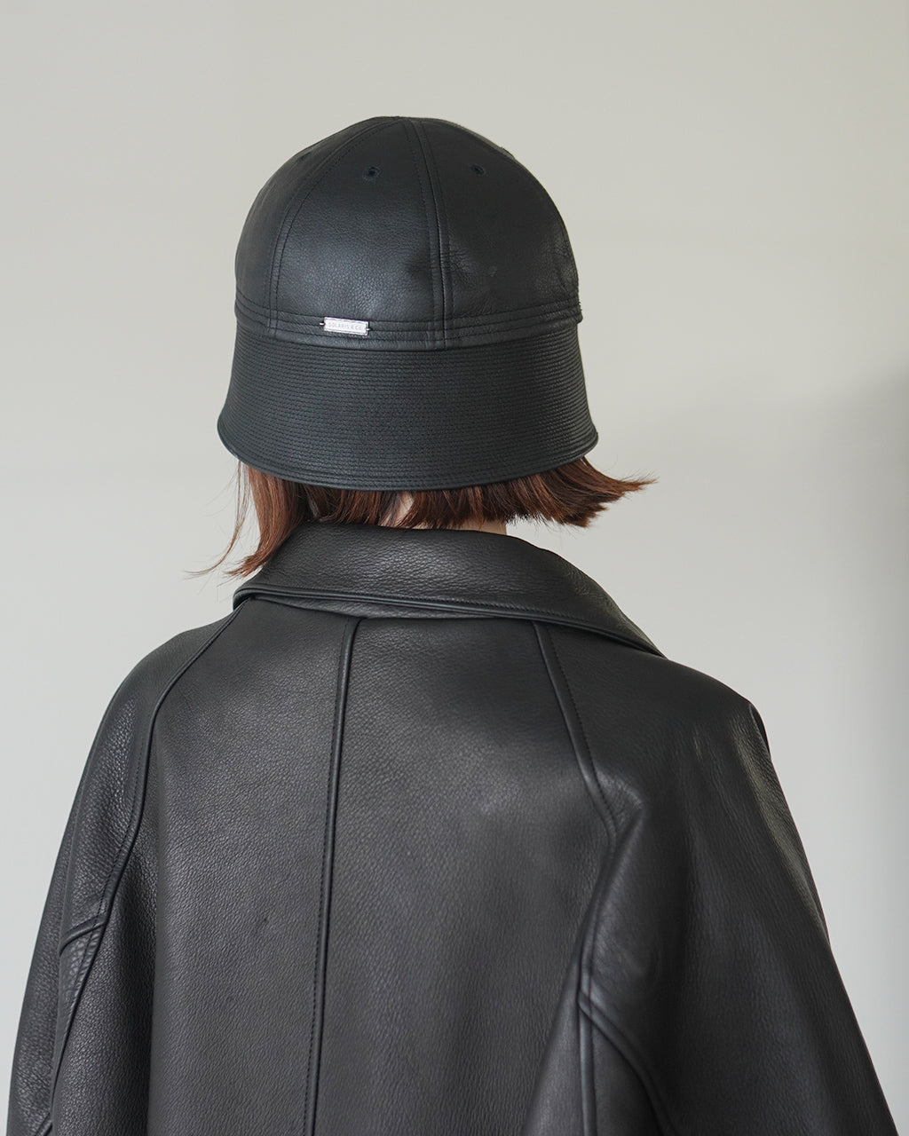 黒のレザーハットを被った人物の後ろ姿。つづれバックルのある帽子の前部が強調され、ジャケットと同素材の質感が映えるショット。