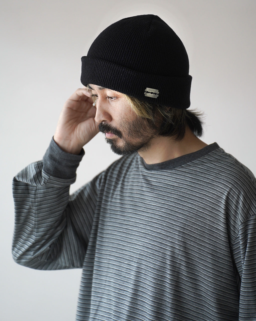 黒のニットキャップ。折り返し部に小型ロゴプレートが付いており、前方に向けて少し斜めに被るスタイル。