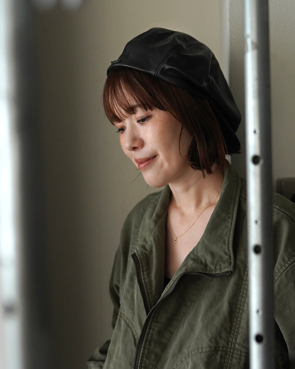 黒いレザー帽子をかぶった女性が、緑のカジュアルジャケットを着用して横向きに微笑んでいる姿。