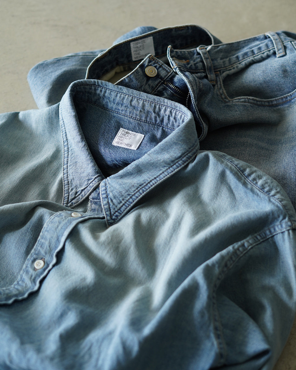 デニムシャツの広めの襟元と胸元のタグが見える、ライトブルーのOVER SIZE DENIM SHIRTのディテールショット。薄い色味のインディゴ風デニム生地の質感が伝わるアップ画像。