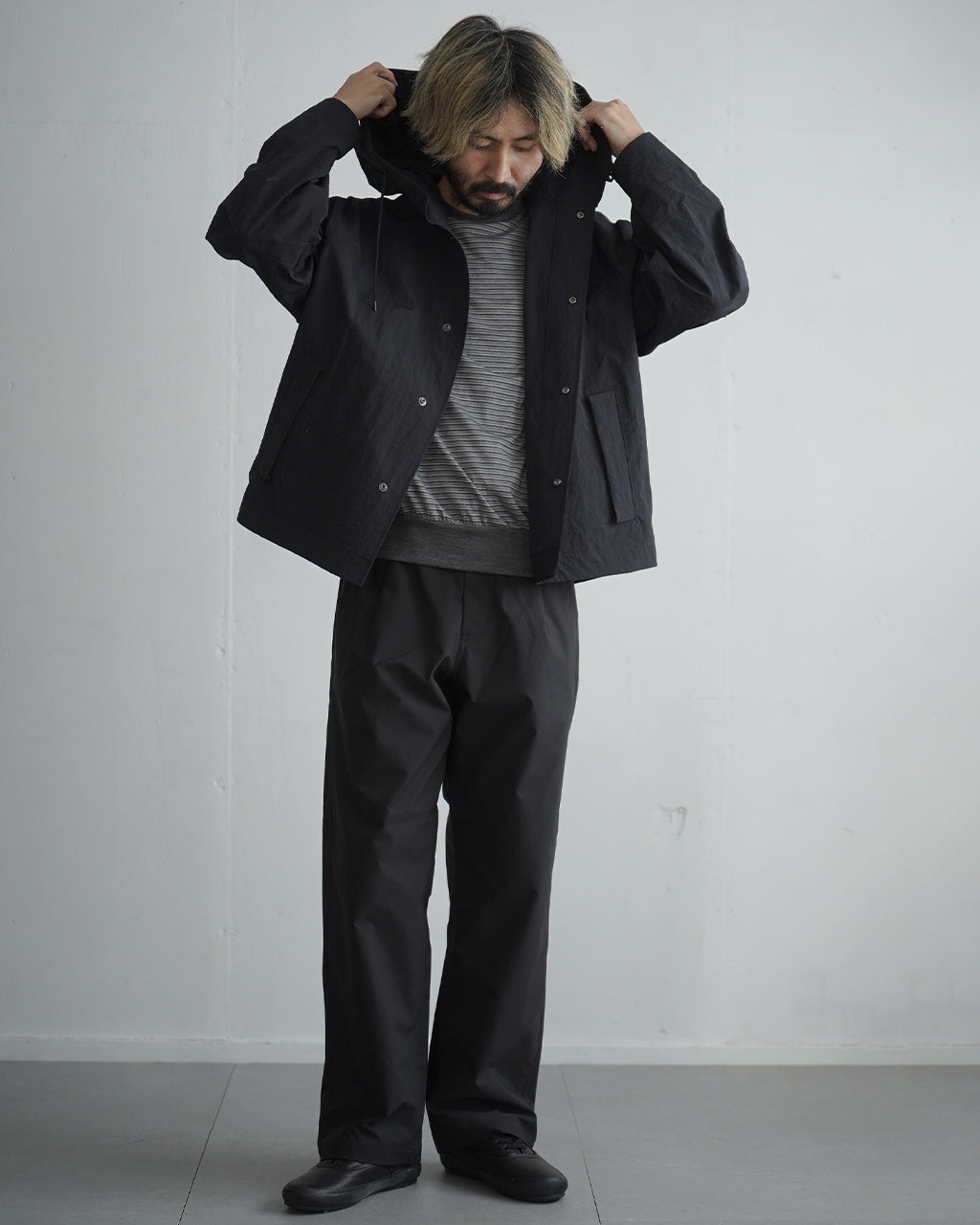 黒系のフード付きブルゾンを着用した男性の全身ショット。撥水性のナイロン生地とゆったりしたシルエットが印象的で、前面のスナップボタンとポケットが見える構図
