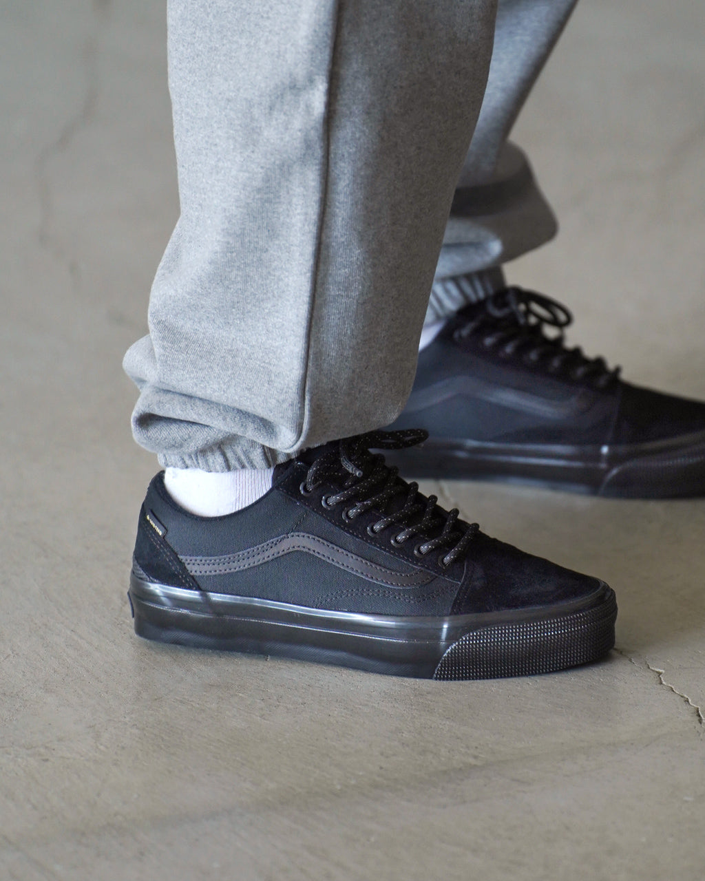 黒系のレザーとナイロンのアッパーにVANSのサイドストライプが際立つローカットスニーカー。ソールはブラック、着用者の脚元の横から見えるスタイル。