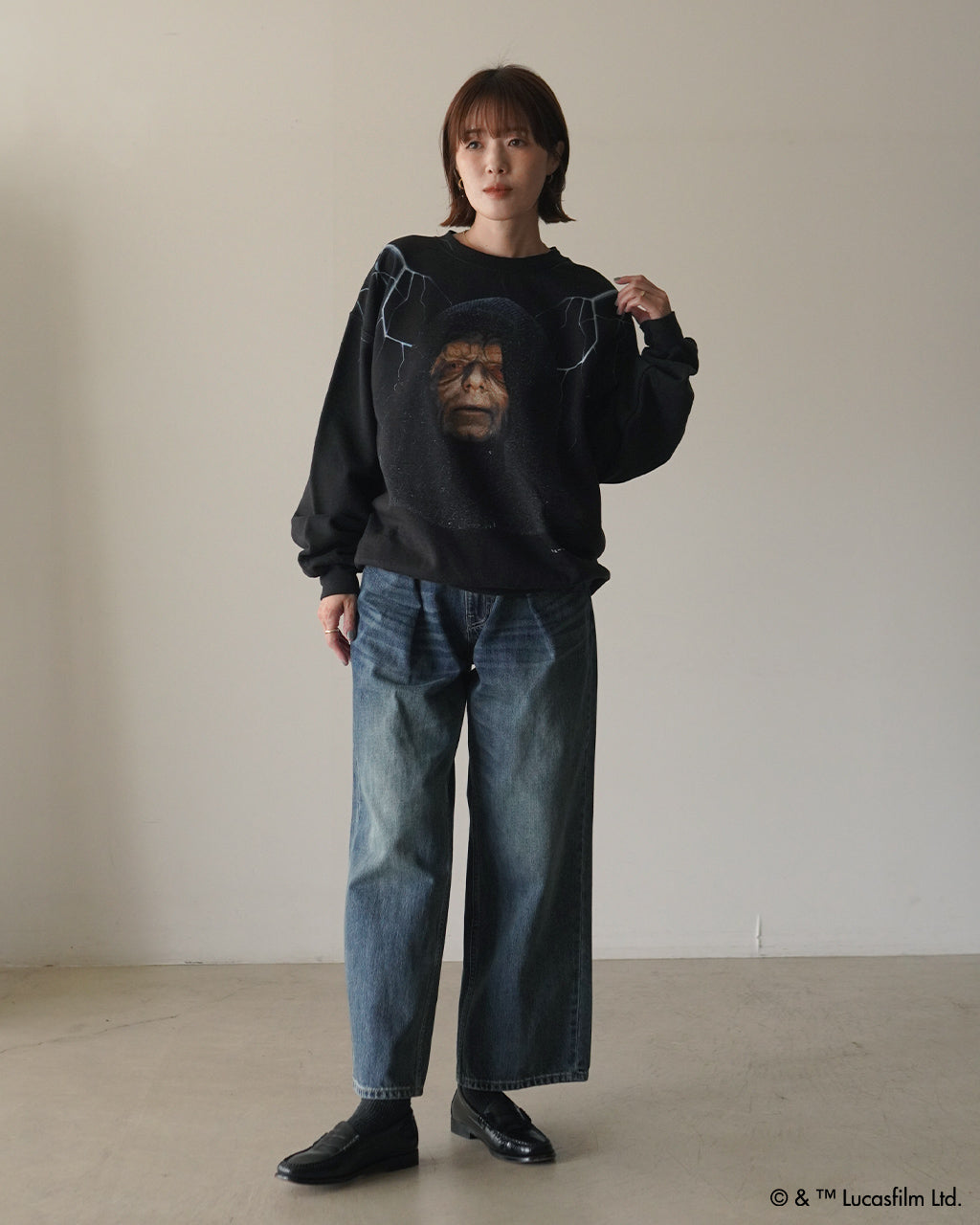 黒い Darth Sidious プリント入り裏起毛スウェットを着用した女性が立つ全身ショット。ゆとりあるシルエットでジーンズとローファーを合わせ、ヴィンテージ感のある表情を演出しています。