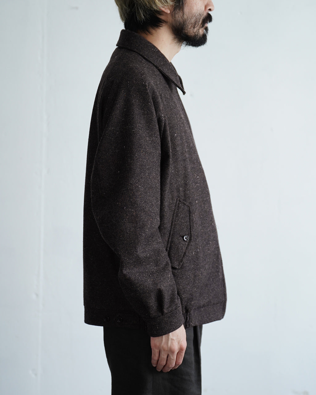 ダークセピアの Harrington ジャケットを横から着用した男性モデル。毛混ジャケットで、サイドポケットと裾のボタンディテールが見えるシンプルなシルエット。
