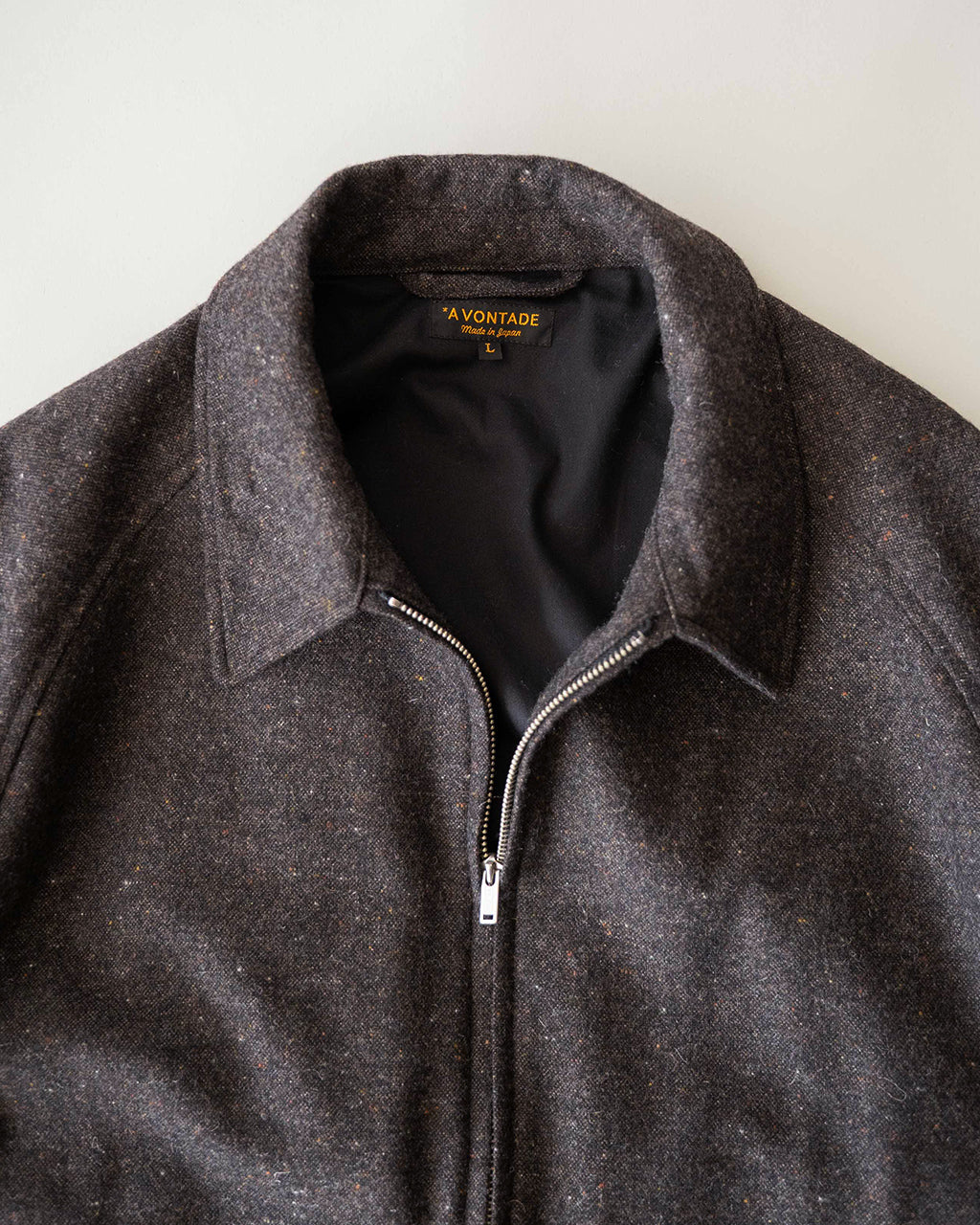 ダークセピア系の Harrington ジャケットのアップ。前立てにジッパーとファスナー、襟は立ち上がった状態で素材はウール混の杢感が見て取れるクオリティ感のある一着。