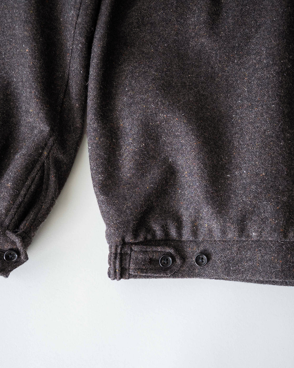 ダークセピア系のウール混素材の Harrington ジャケットの袖口と裾付近をアップで撮影。生地の杢感とボタン留めのディテールが見える