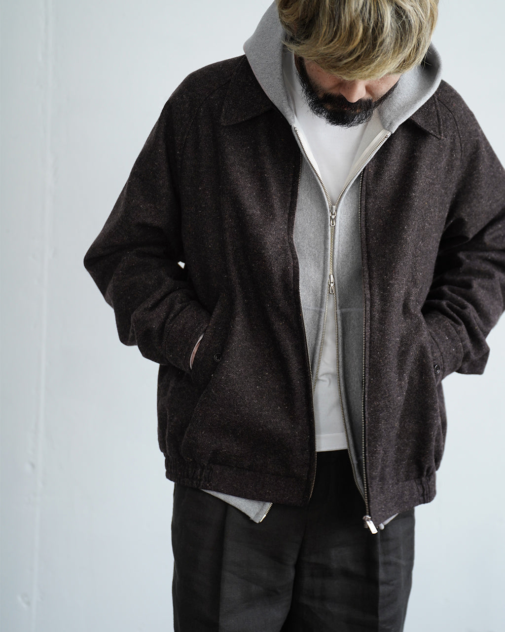 ヘッドを下げた男性が暗い杢調の Harrington ジャケットを前ボタンとジップで着用している姿。内側はグレーのフリース/ライナーが覗き、裾と袖にはアジャスターが見える。