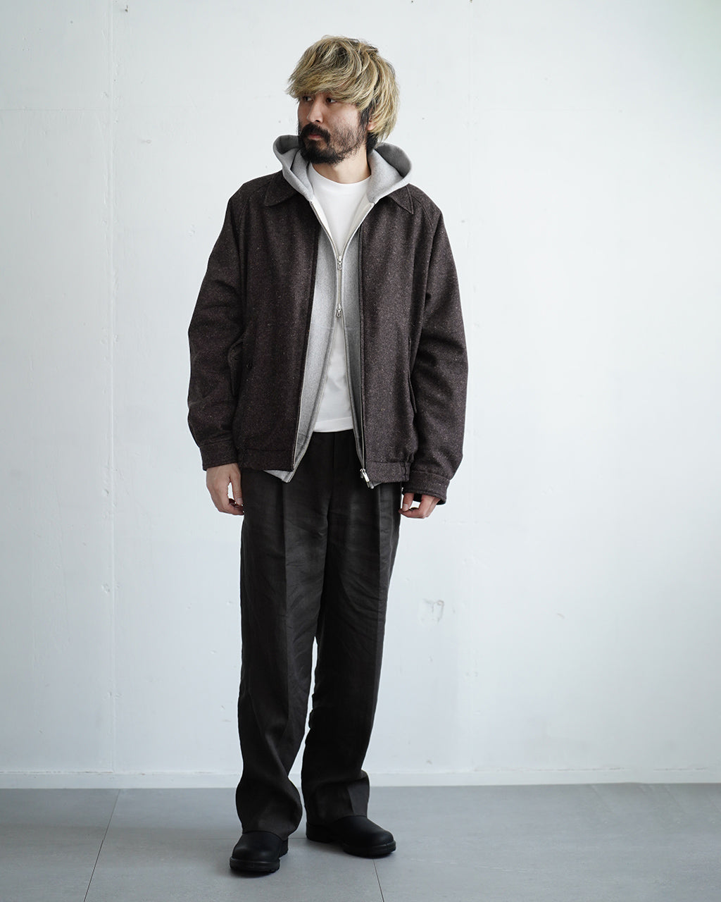 ダークセピアの Harrington ジャケットを着用した男性モデルの全身ショット。前を開けた状態で、内側に白いインナーとグレーのフード付きライナーが覗くデザイン。