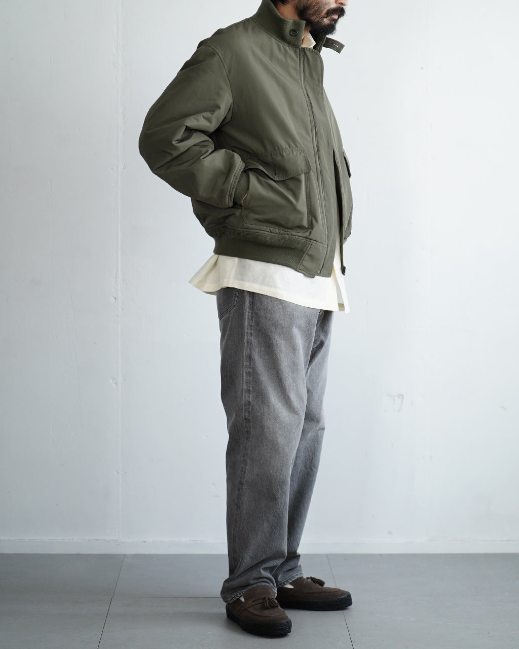 olive系のPadded Wep Jacketを着用した男性の横からのシルエット。ファスナーとリブのディテール、前ポケットの膨らみ、裾の絞りが目立つスタイル。裾からのレイヤードで白シャツの裾が見える着用イメージ。