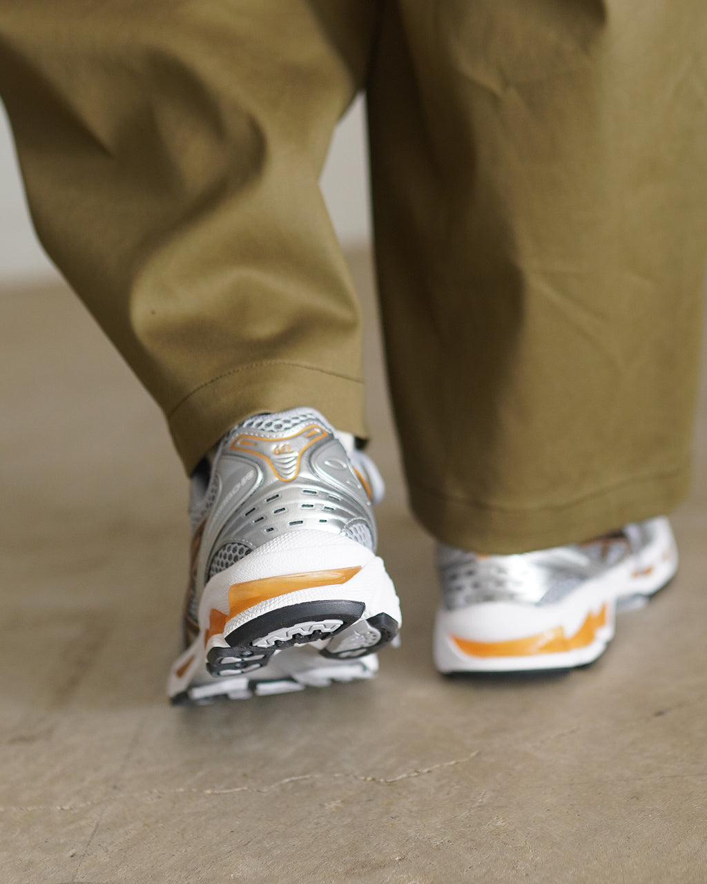 シルバーを基調としたASICS SportStyle GEL-KAYANO 14の後方斜め視点。ゴールド・ブラックのラインがアクセントになり、厚めのソールとゲルクッション風の装飾が特徴的なランニングシューズの背面と踵部分が見える。