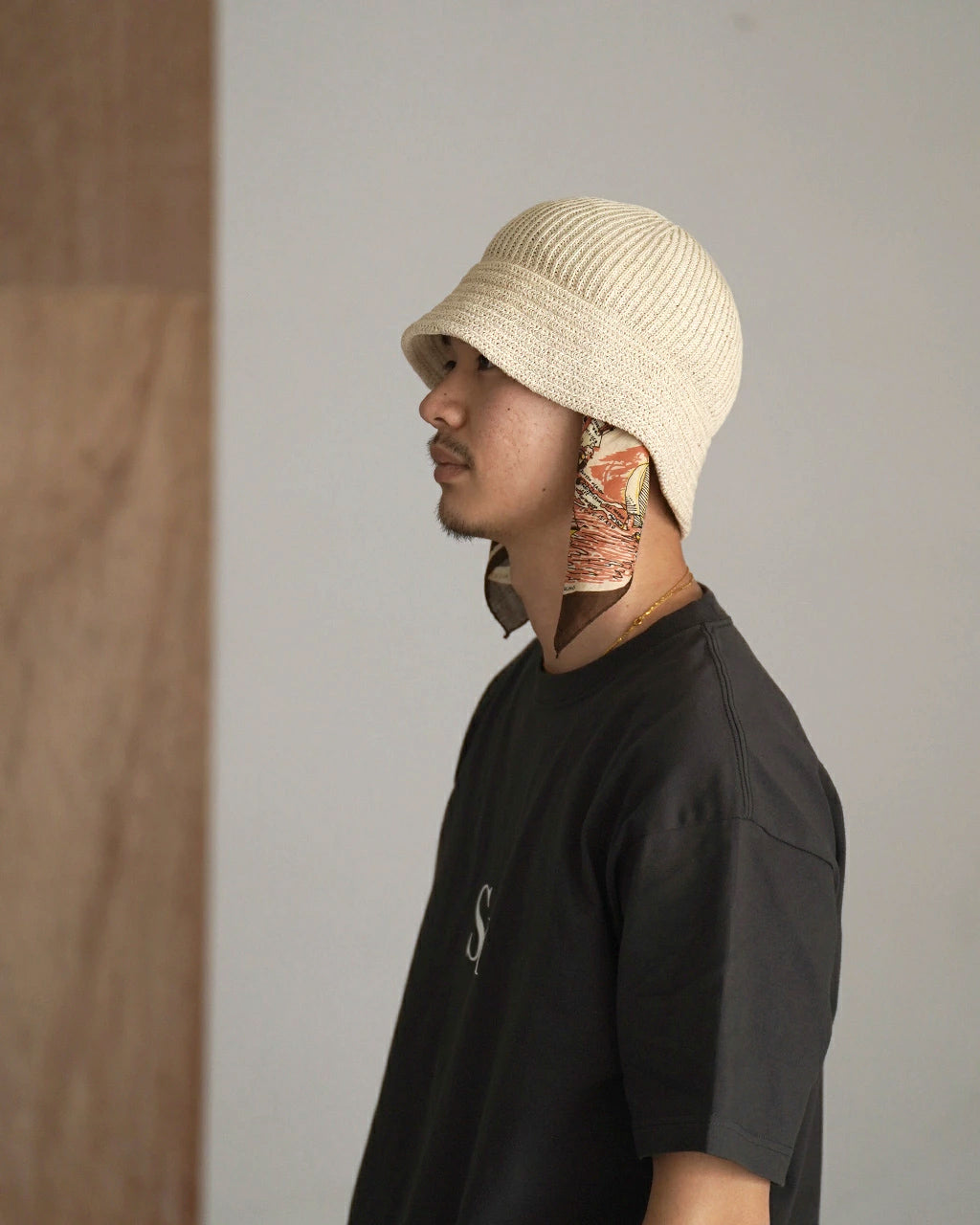 ベージュの編み地キャップに布製スカーフを耳の後ろで結んだ男性の side view。ダークTシャツと落ち着いた背景。