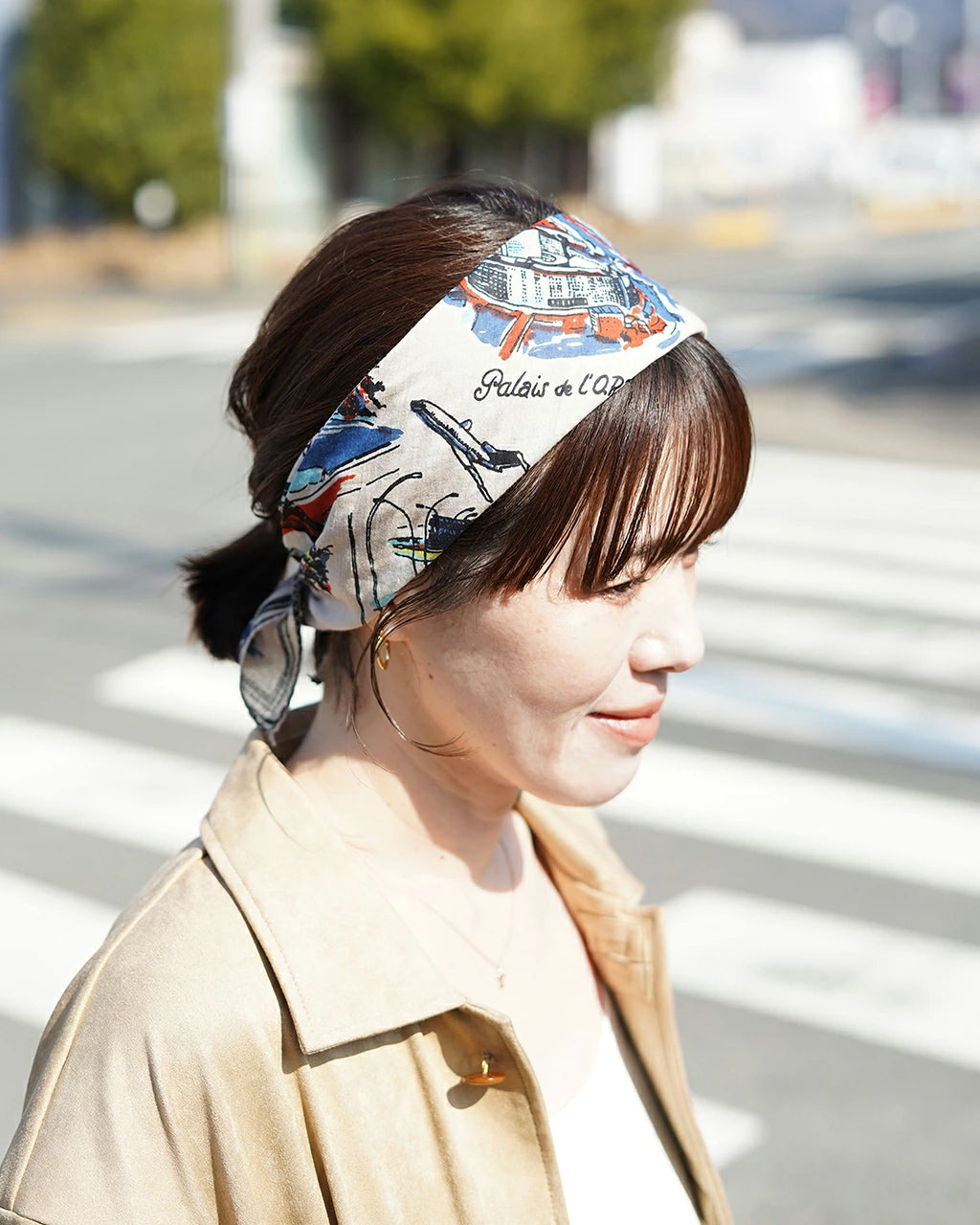 白地に柄入りのバンダナを頭に巻いた女性の横顔。都市の路上を背景に光が差し、柄には船や建物のモチーフが描かれている