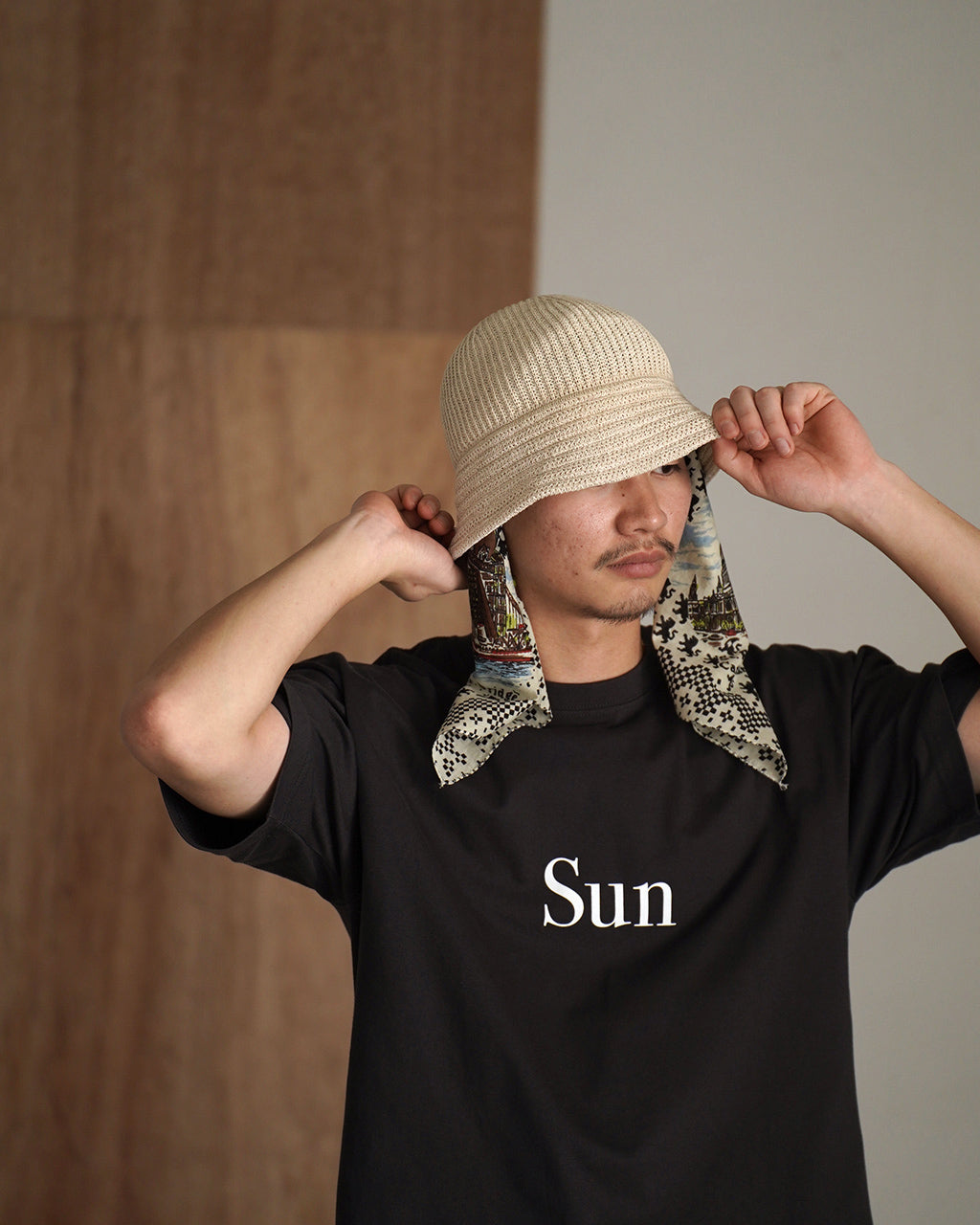 ベージュの編み地ハットを被り、黒のTシャツに長い柄スカーフを取り付けた男性モデルの写真。前方を見据え、カジュアルな雰囲気。