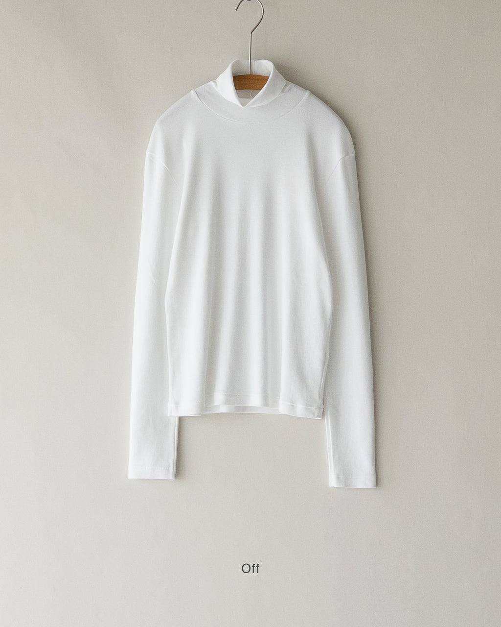 白のタートルネック長袖Tシャツがハンガーに掛かって壁に掛けられている様子。光沢を抑えた素材感とゆったりとしたシルエットが特徴的。