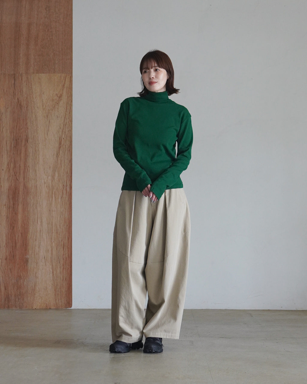 ダークグリーンのタートルネック長袖Tシャツを着た女性が、ベージュのワイドパンツを履き立っている様子。裾まで落ち着いた印象のシルエットとリブ編みの生地感が際立つ。