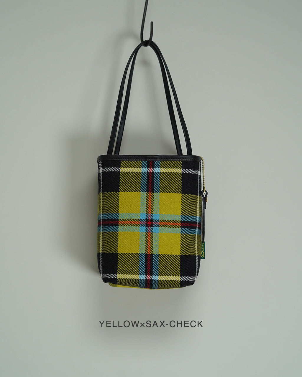 Yellow×Sax-Checkのタータン柄ショルダーバッグ、黒いレザーハンドルとジッパー付きの巾着型デザイン。前面にチェック柄が全面に表れ、控えめな縦長シルエットが特徴です。