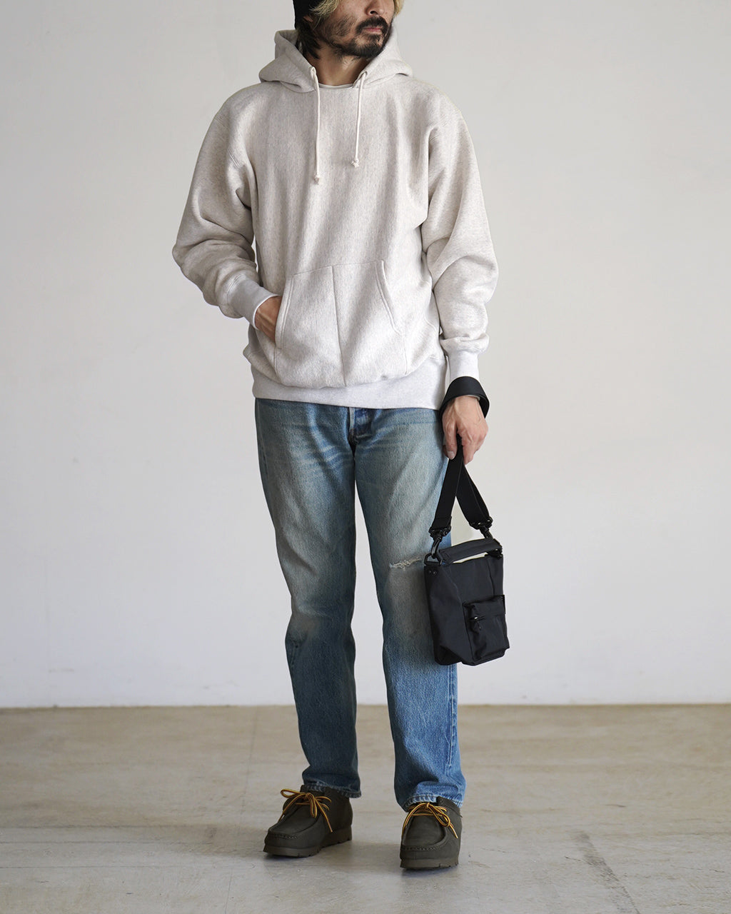 淡いグレーのフリースパーカを着た男性の全身写真。フード付き、前面ポケットが目立ち、 jeans に合わせたカジュアルなスタイルが映える。小物として黒のショルダーバッグを手に持つ。