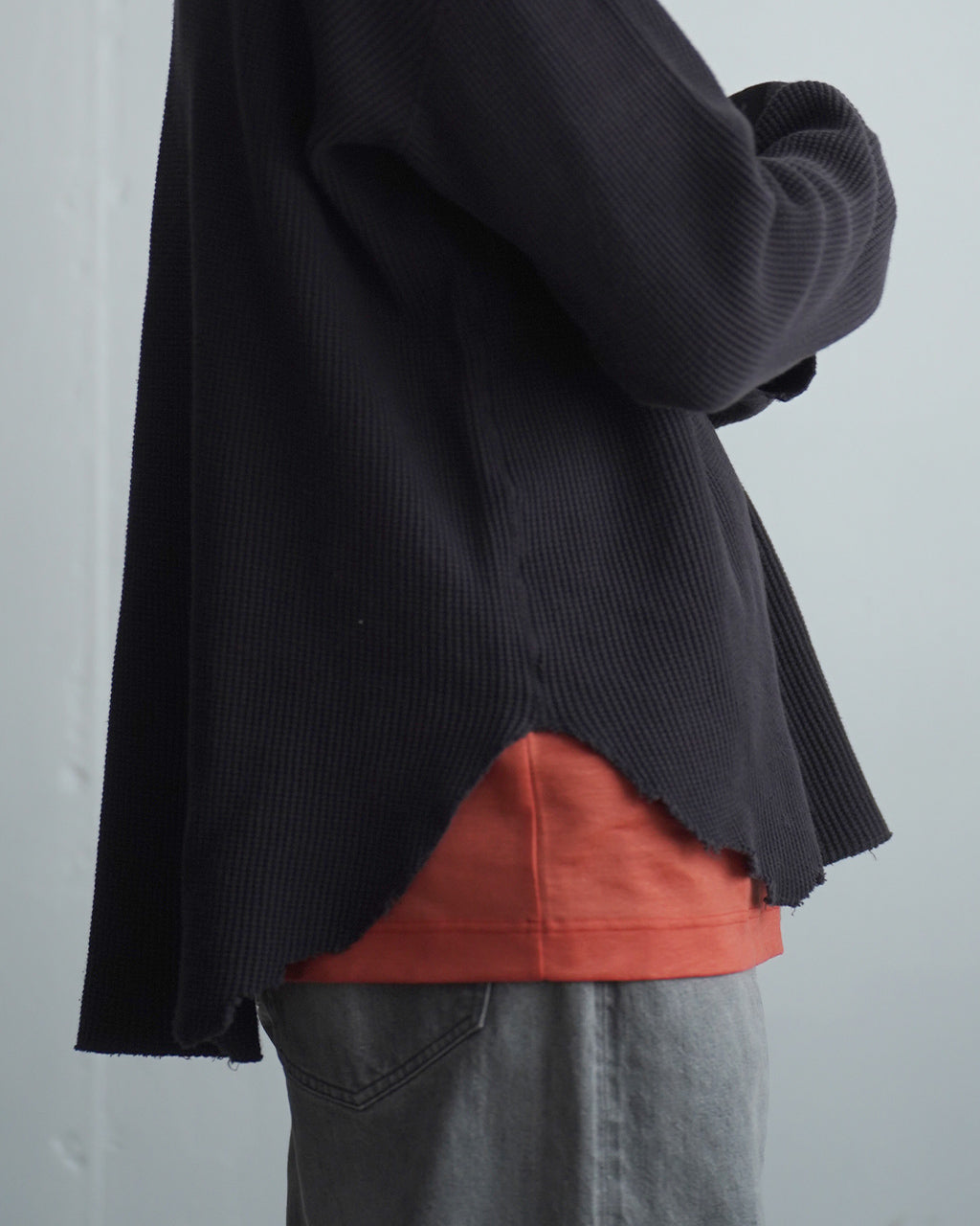 黒系ワッフル素材の長袖Tシャツを着用した人の横からのシルエット。裾はラウンドカットで裾の赤系シャツが覗く。凹凸のある生地感が特徴的。