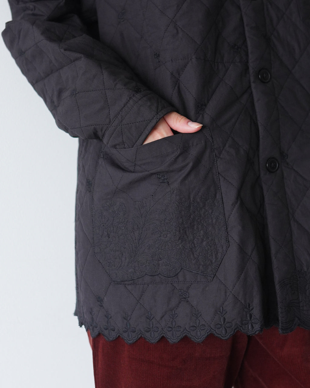 黒系の刺繍入りキルティングジャケット。前立てのボタンと袖口、裾にかけて広がるスカラップレース風の装飾が特徴的で、ポケットに手を入れる様子が映る。
