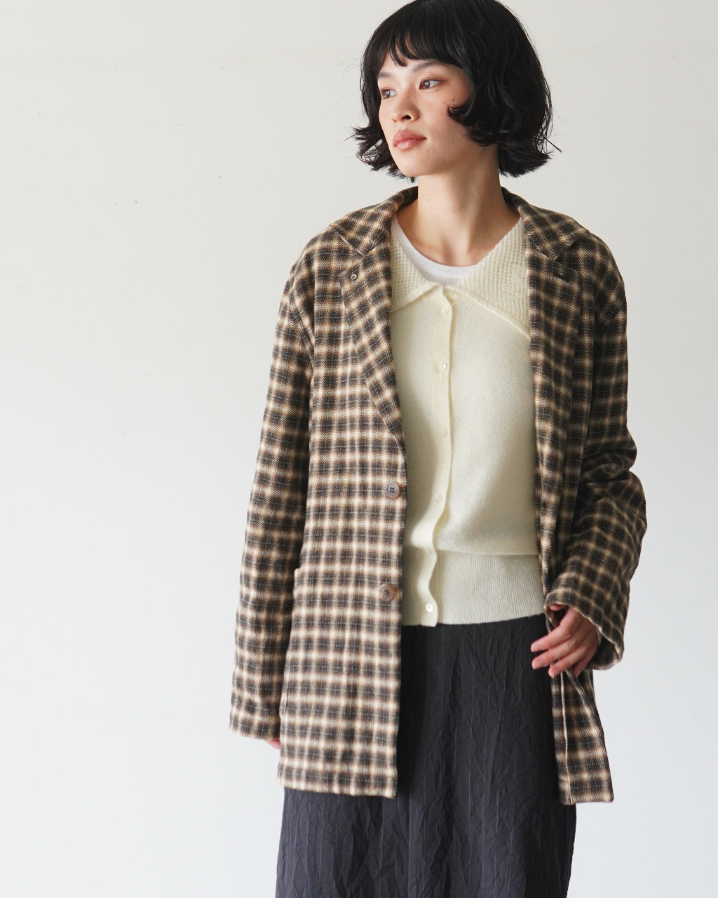 グレー系チェック柄のジャケットを羽織る女性モデルの正面寄りショット。肩口まで広がる襟付きデザインと落ち着いた色味が特徴。