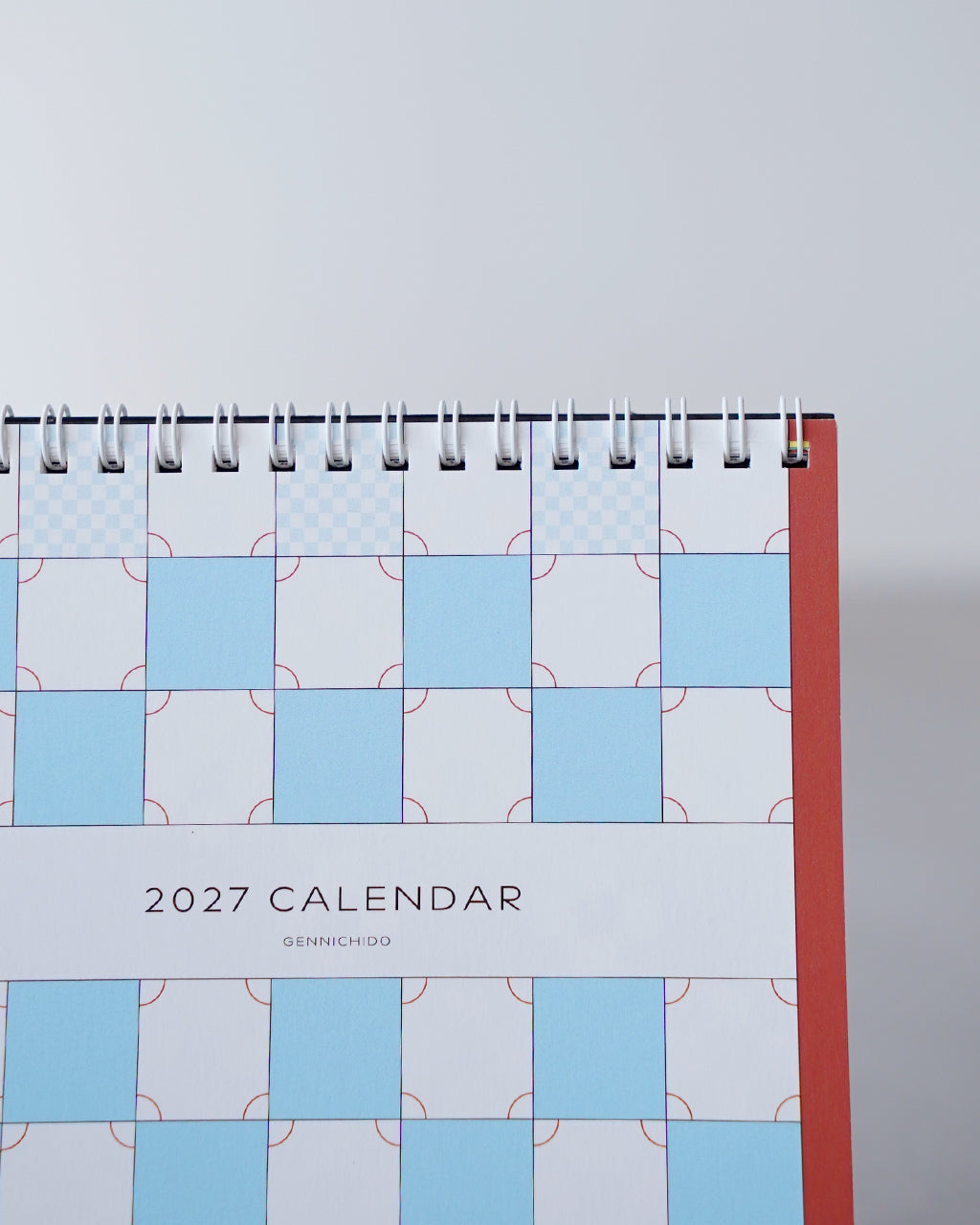 チェス柄の卓上カレンダーの表紙アップ。白地に青と水色の市松模様と黒文字の2027 CALENDAR、背面は赤い台座の一部が見える。
