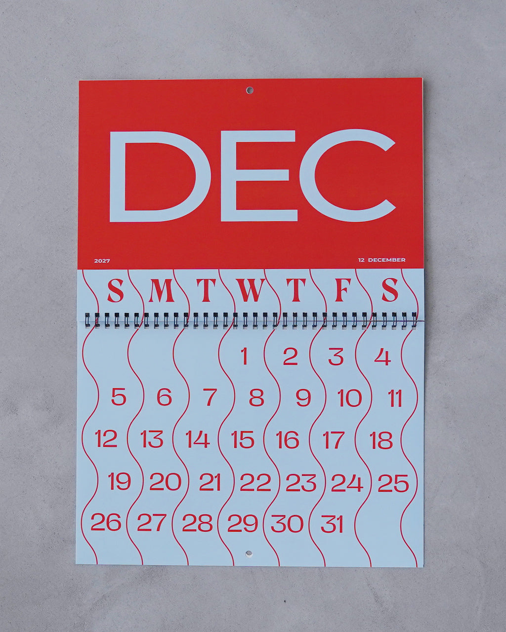 赤と白の大きな文字が印象的な2027年用リング綴じカレンダー。上部の赤い面に「DEC」と大胆に配置され、下部には月日が波形の模様とともに並ぶデザインです。