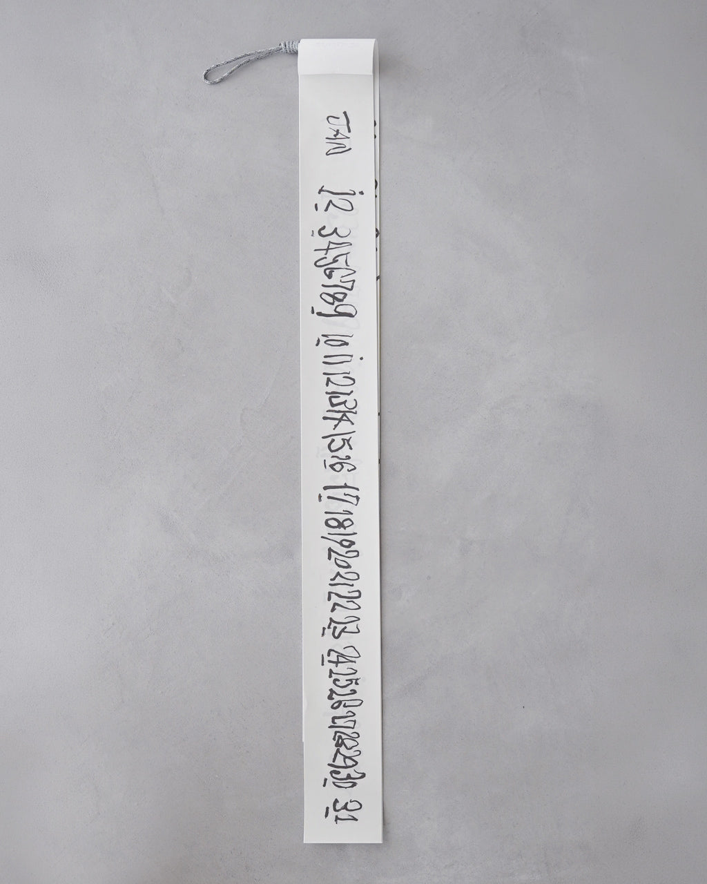 細長い白い帯状のカレンダーが垂直に写り、紙の両端に黒い手書きの数字が連ねられています。ストラップが左上部に付いたデザインです。