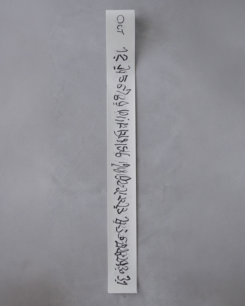 白い長尺の紙に手書き風の黒文字が縦書きで並ぶ垂直デザイン。背景は無地の灰色壁で、細長い形状のカレンダー用紙が画面中央を縦に伸びている様子。