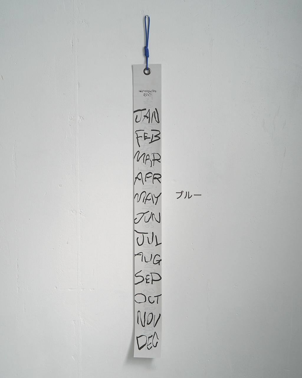 白い細長い縦型カレンダーに、月名が黒抜きの手描き風文字で並ぶ。上部に紐付き、背景は淡いグレーの壁。日付をめくる短冊型デザインが印象的な風景写真。