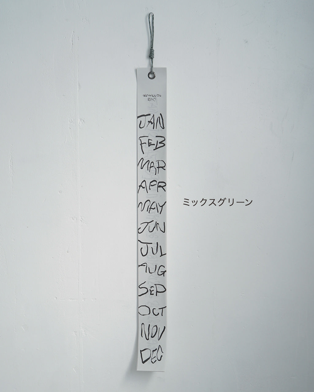 縦長の白い短冊カレンダーが壁掛け用紐で吊るされ、月名が手描き風で連なるデザイン。背の高いスタイルで、表紙にもブランド名の文字が小さく入る。