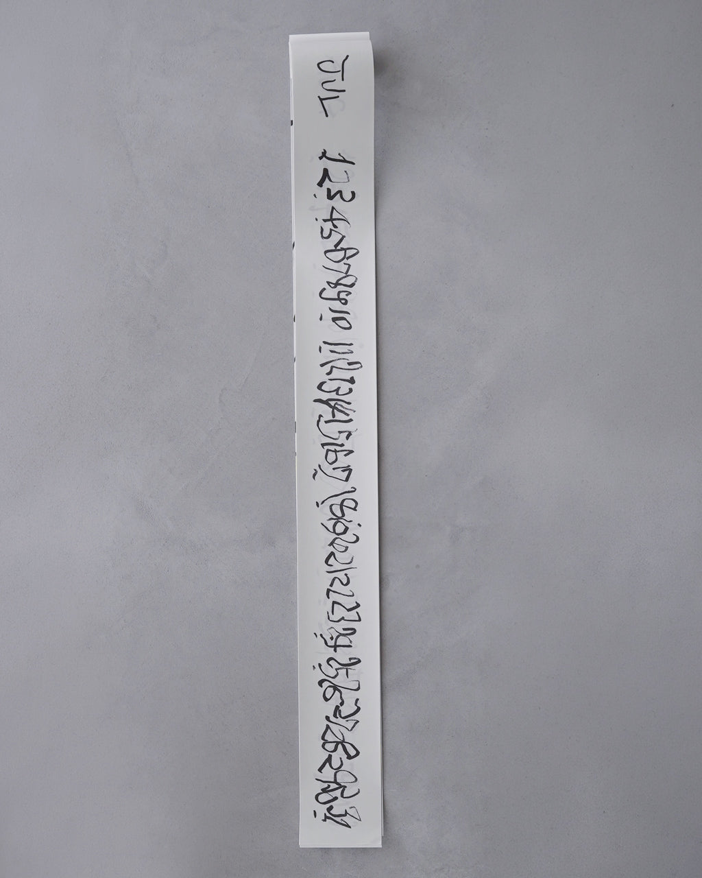 縦長の短冊状カレンダーが壁に掛かり、黒の手書き風数字が連なるデザイン。白地に描かれた文字が目を引く、AURA Tanzaku Calendar の表紙が露出している画像です。