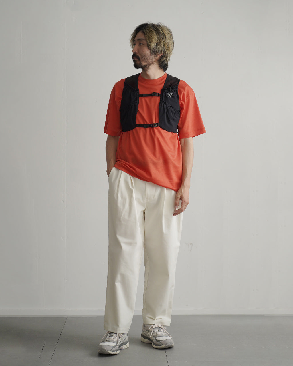 赤色の半袖Tシャツに黒のベスト型ベストストラップを重ね着した男性モデルの全身写真。ベストは胸元のロゴと前後でポケット付きのデザイン。背景は淡い壁面で、動作を想定した装着イメージ。