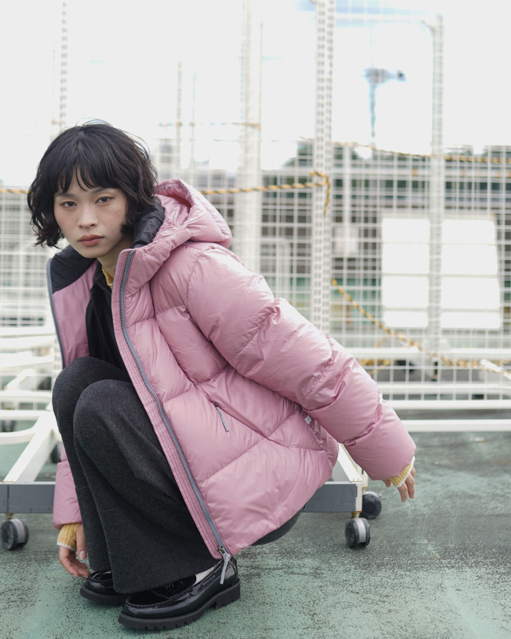 ピンクのダウンジャケットを着た女性が屋外で座り込む姿。ショート丈で裾まで暖かそうなボリューム感とフロントのジップ、立ち上がるフードが特徴的です。