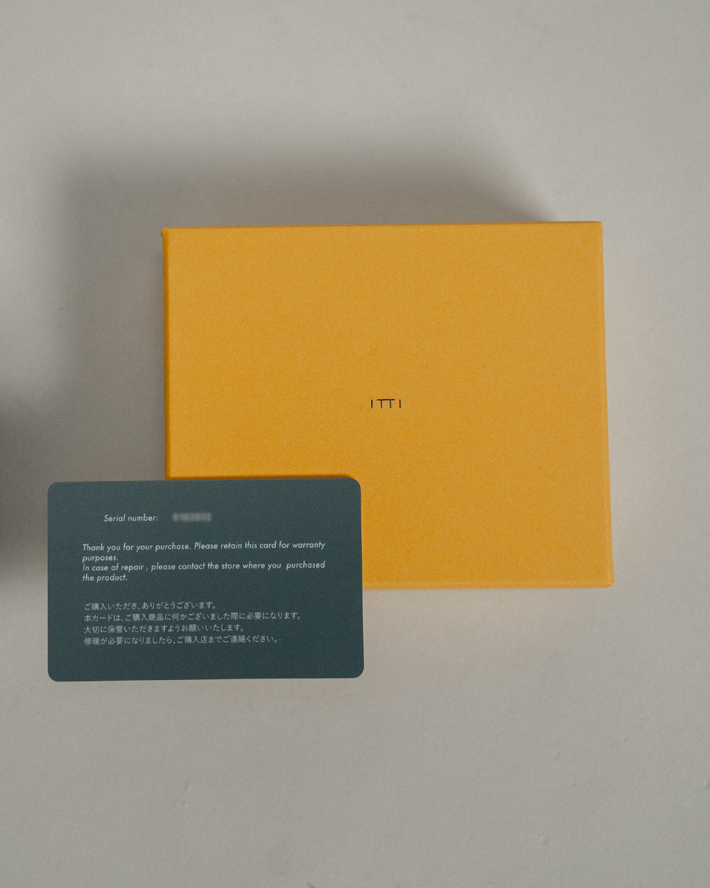 イッチ ITTI CRISTY WIND COMPACT WLT クロコダイルレザーの黒いミニウォレットと付属カード。鮮やかなオレンジの箱とロゴが中央に配置された写真