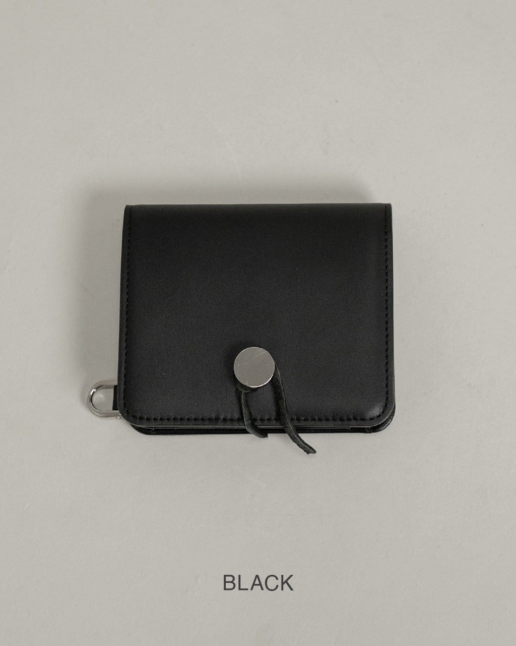 ブラックカラーの小型財布。正面に丸型の金具と紐の留め具が目を引くデザインで、コンパクトなシルエットが特徴。厚みは抑えられ、持ち運びやすいサイズ感が伝わる一枚。