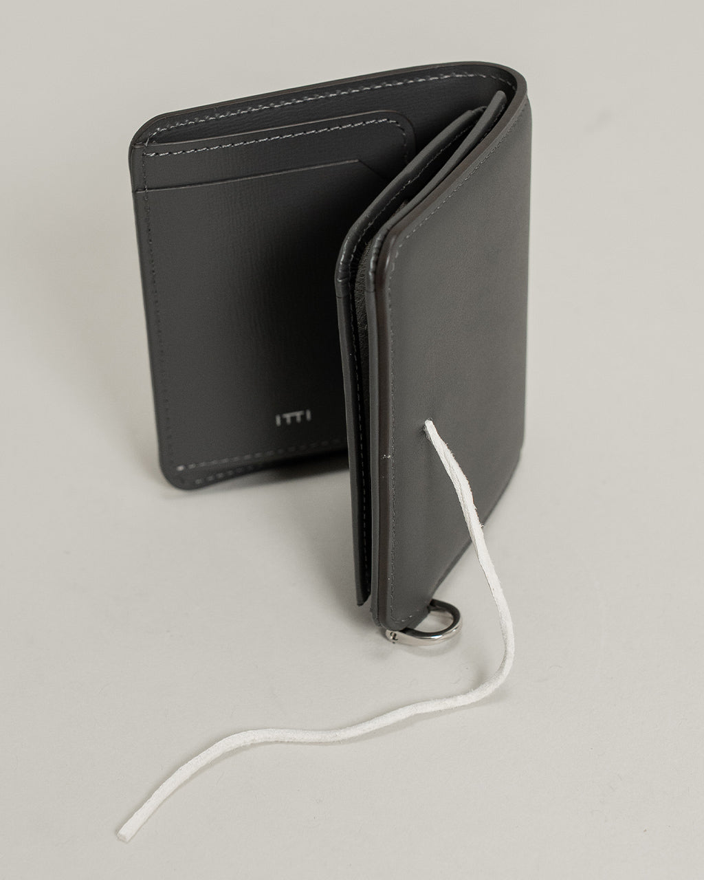 ブラックのミニマムな折り財布。内ポケットとステッチが際立ち、Dカン付きの金具と白い紐がアクセントとして見える雅やかなデザインです。
