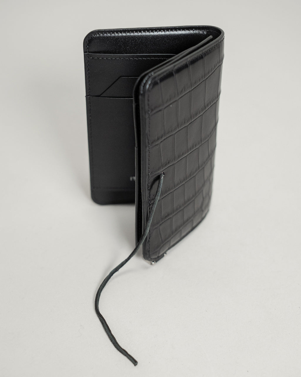 黒のクロコダイル型押しのミニウォレット。背面と側面の立体感が伝わる、縦長のコンパクトなデザイン。紐を結んだディテールがアクセントとして見える。