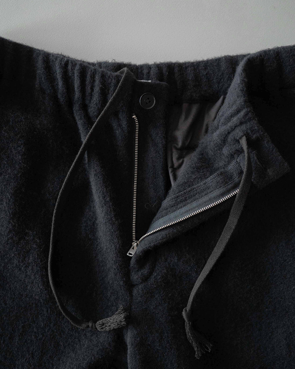 ブラック系のSilk Wool Fleece Pantsのファスナー付き前身ごろアップ。ジップと裾を絞る紐、ウエストゴムのデザインが見える正面ディテール。