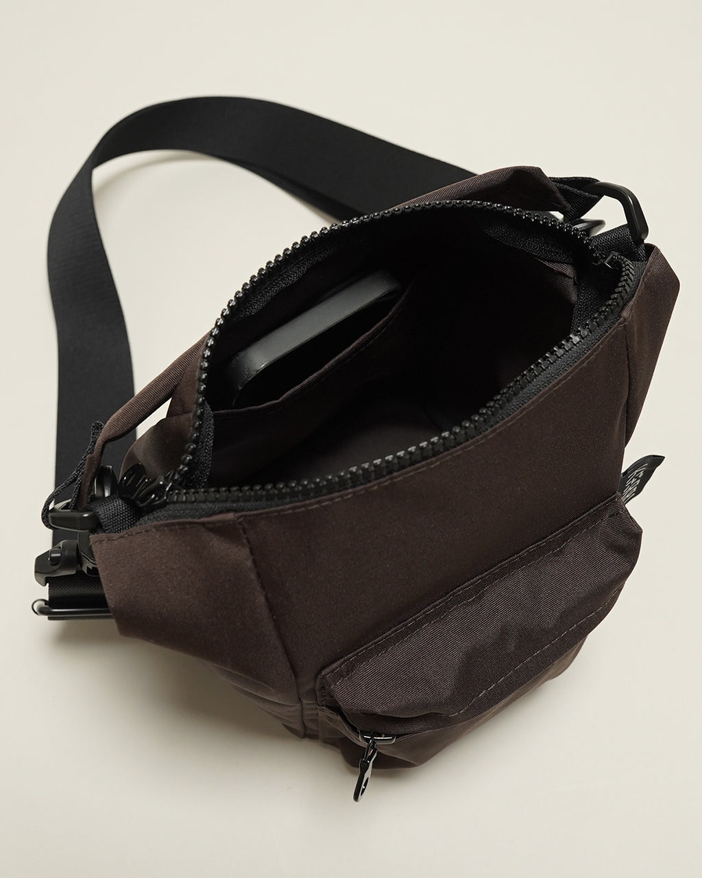 ダークブラウンの2WAYショルダーバッグ。主室はジップ開閉で中にスマホを入れたカーセルが見える。ショルダーストラップと外ポケットが特徴的。