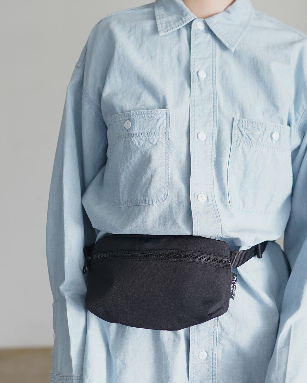 KARRI AMIACALVAのWAIST BAG K011。青系のデニムシャツと合わせた着用イメージ。前ポケット付きの小型ウエストバッグが腰に装着された状態で、ブラックの本体とファスナーが際立つデザインです。