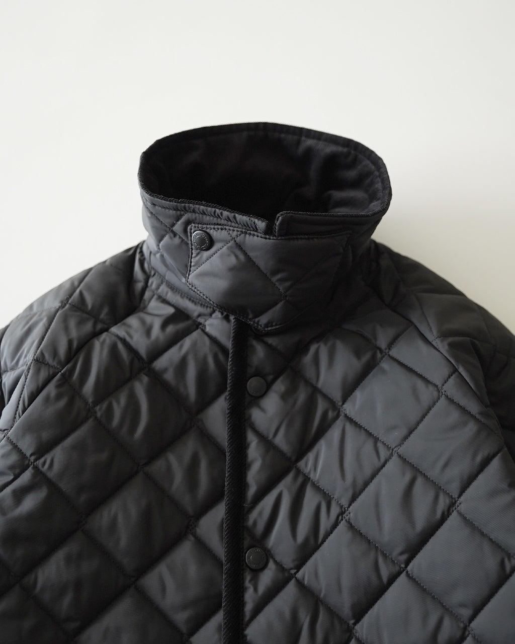 ブラックのキルティングジャケットのアップ。高めのスタンドカラーとダイヤモンドキルティングのパターンが特徴的。前立てはボタン留めで、首元のチンストラップが見えるディテール画像です。