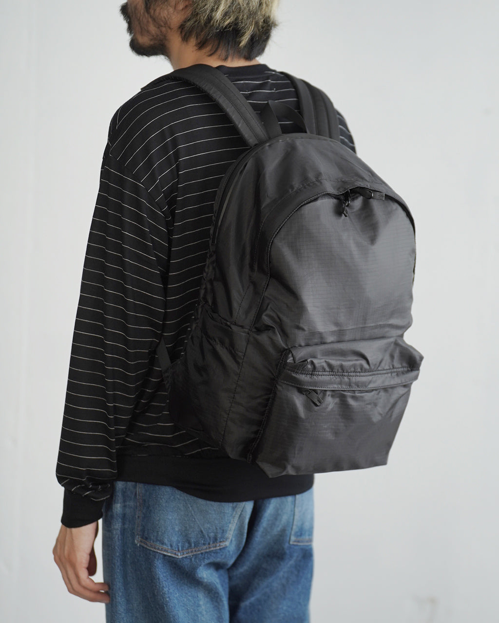 LC by LOWERCASE の DAYPACK LITE 20Lを背負ったモデルの後ろ姿。黒のストライプ長袖トップスとデニムパンツを合わせ、軽量でシンプルな黒色リュックが目を引く