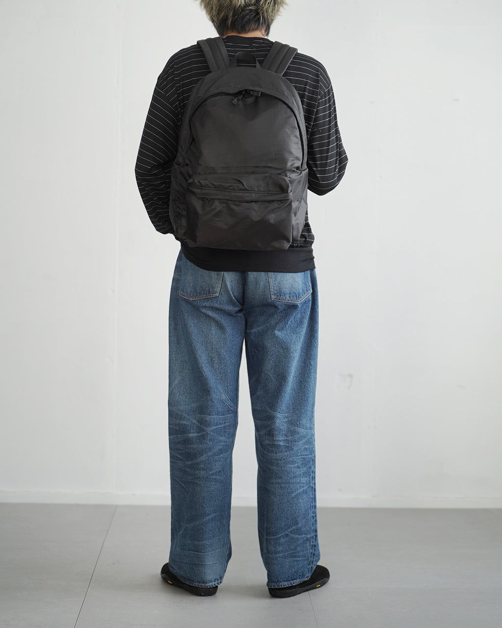 背面から見る黒の LC DAYPACK LITE 20L、肩紐と本体にナイロン素材、ジーンズと黒の長袖トップスを着用した人物が背負っている様子