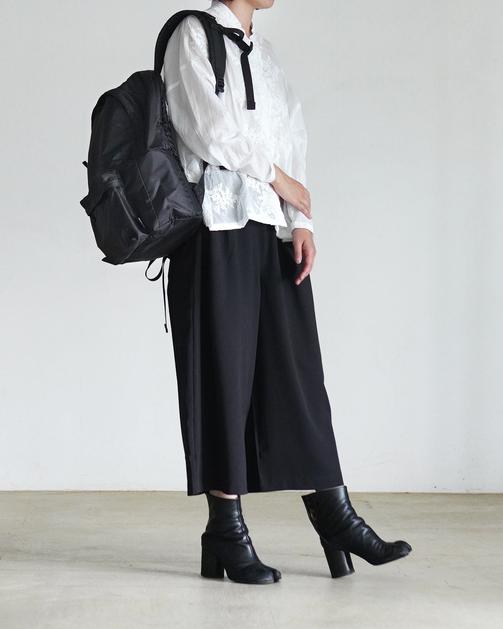 黒のリュックを背負ったモデルが白いトップスと黒のパンツに合わせ、長めのブーツと共に前方左横から撮影された着用ディテール写真