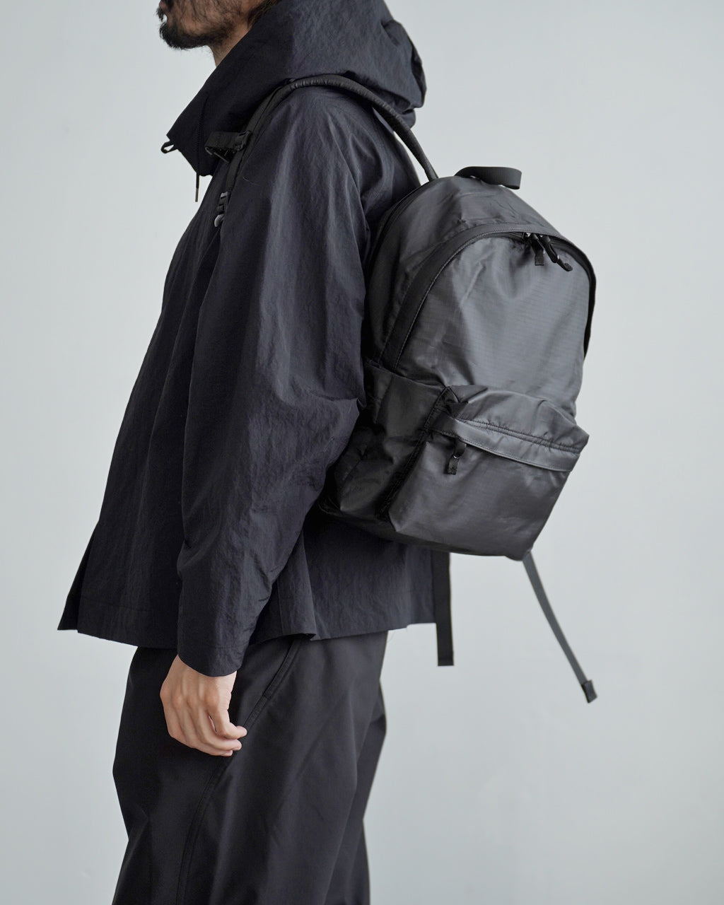 LC by LOWERCASEの DAYPACK LITE 16Lを背負った男性の横顔とバックパックの全体像。ダークトーンのジャケットと同色の軽量バックパックで、左側にファスナー付きポケットが見える。