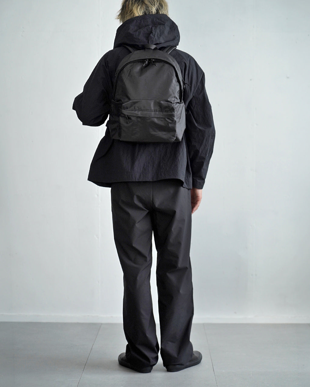 黒系のLC by LOWERCASEのバックパックを背負う人物の後ろ姿。丸型のジップ付き前ポケットと丈夫そうなリュック本体が特徴。全身黒で統一されたアウトドア系スタイルを披露。