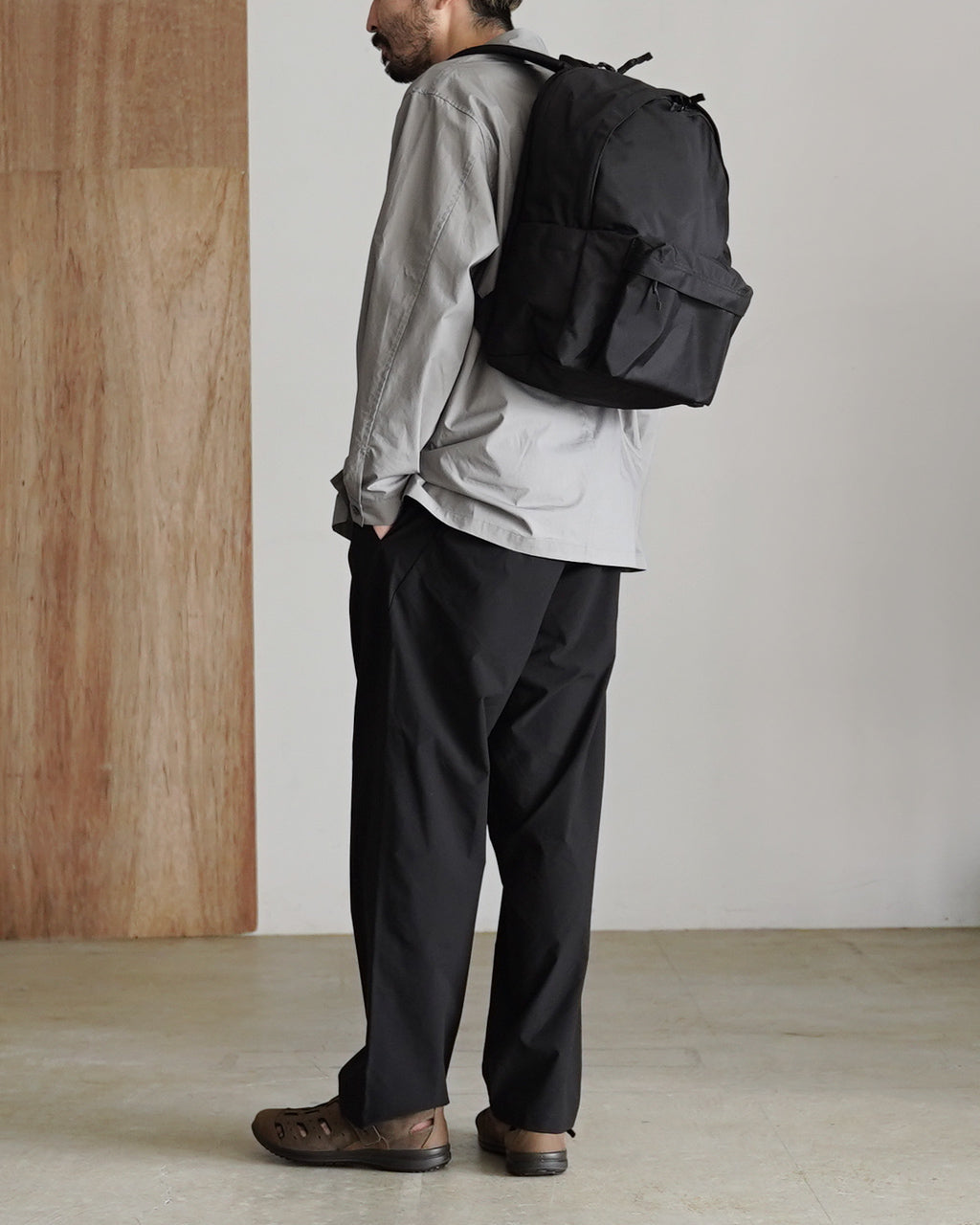 グレーの長袖シャツを着た男性が黒の DAYPACK Msize を背負っている様子。背面ショットで、背中側の荷物が目立つブラックリュックの全体像が確認できる