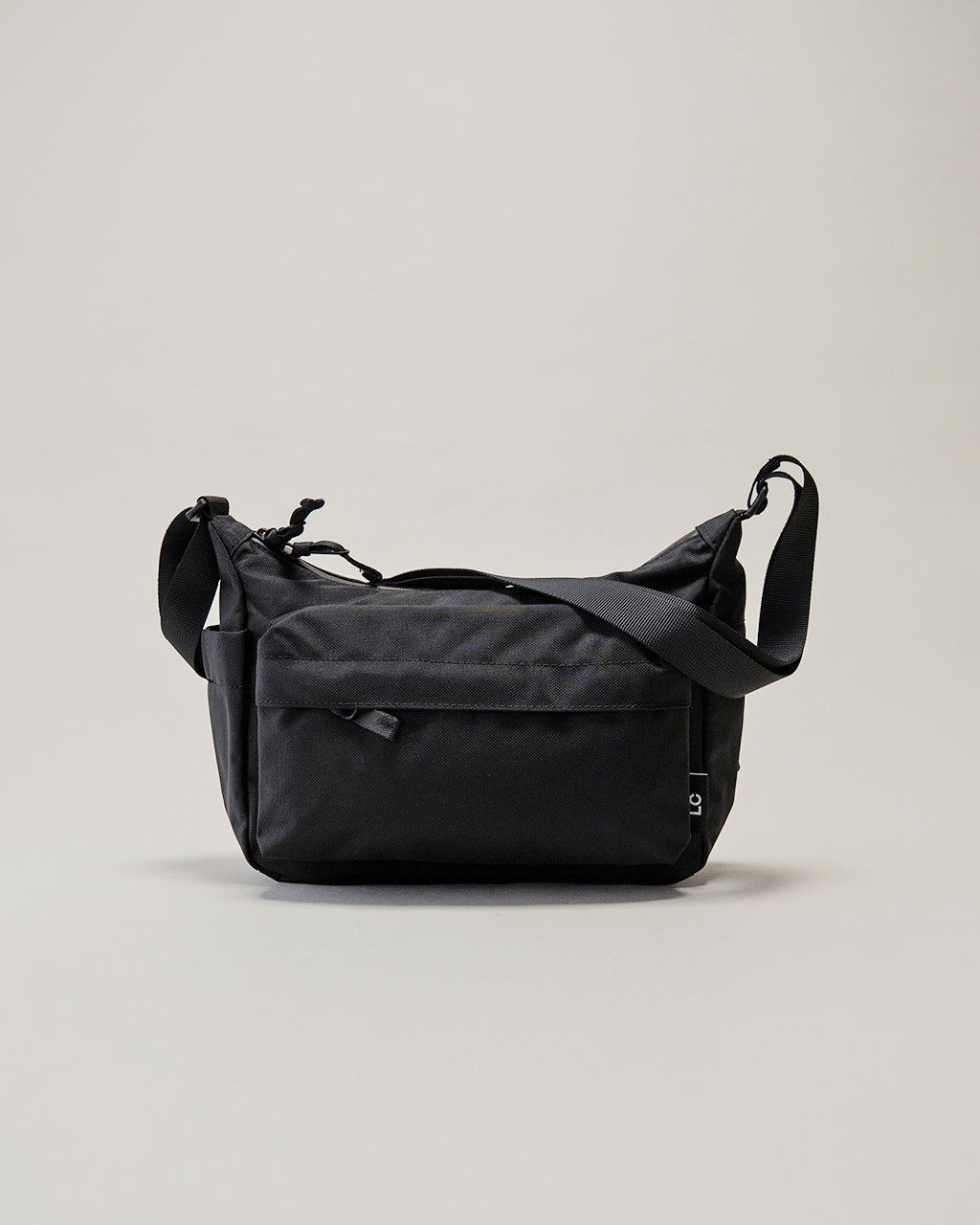 LC by LOWERCASEのショルダーバッグ、前面にはジップ付きポケットが見えるブラック系のシンプルなデザイン。全体はナイロン素材で、長めのストラップが配置された横から斜め下方へ垂れる姿。