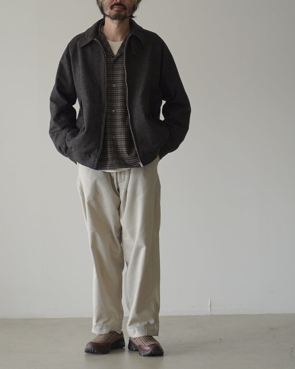 ブラック系のジャケットを着た男性の全身写真。内側にチェックシャツと白Tを重ね着したカジュアルなスタイルで、落ち着いた雰囲気のコーディネートを示す。